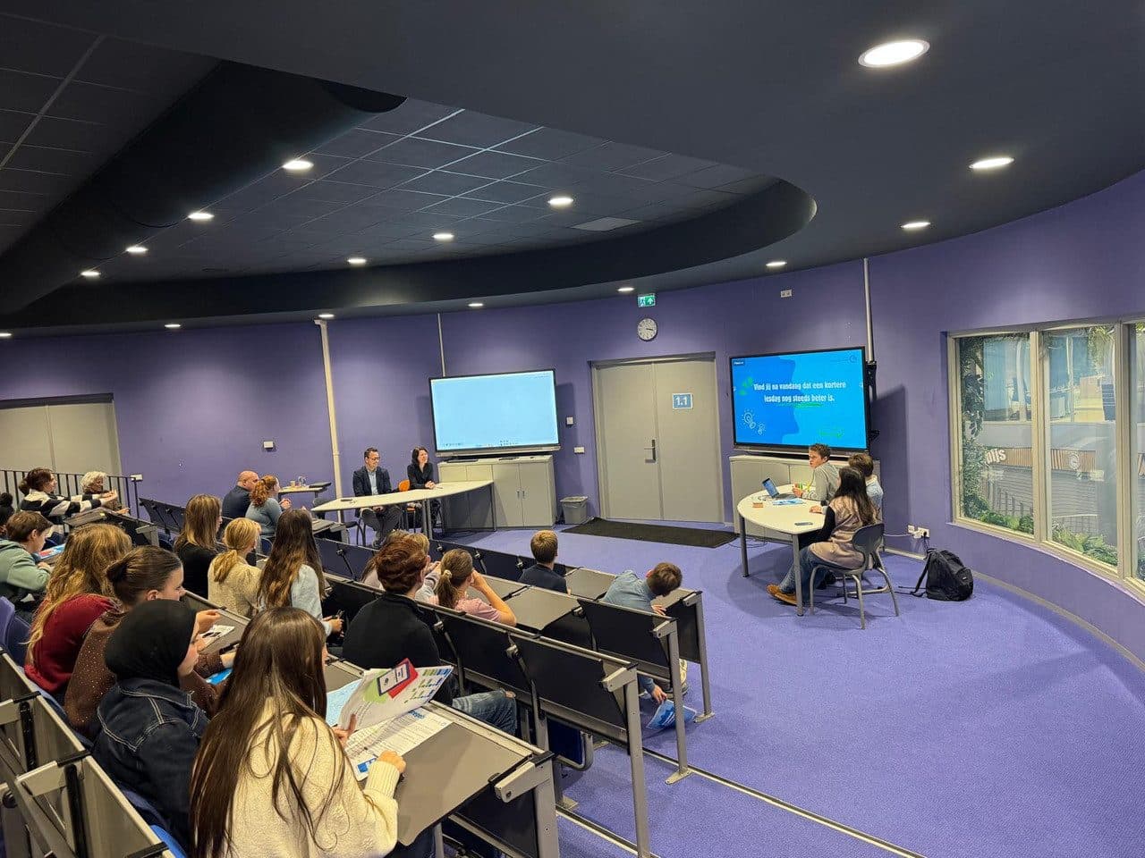 Een volle collegezaal met leerlingen, met vooraan een paneltafel en een presentatie op het scherm