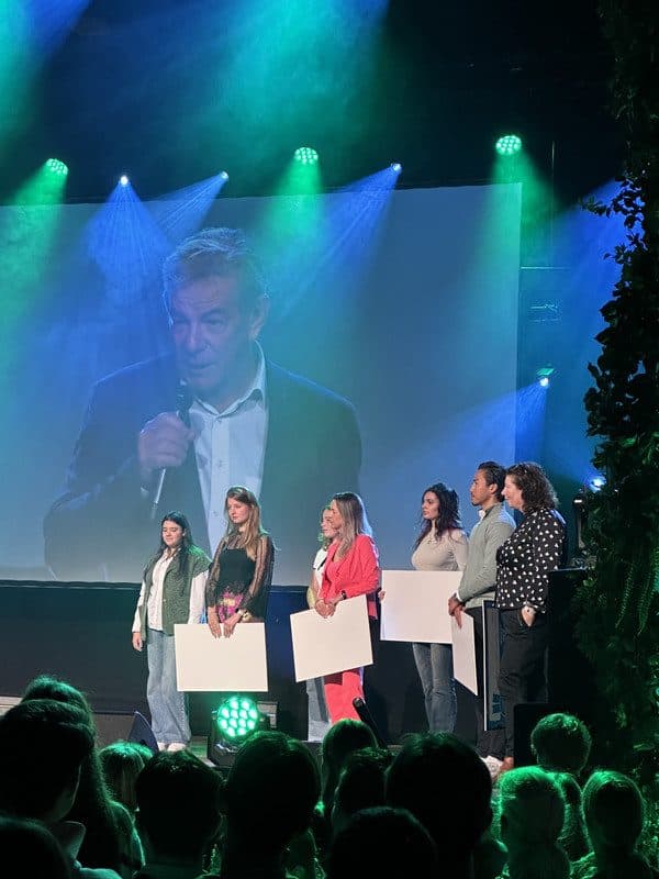 Een podium met sprekers en deelnemers, met een groot scherm en groen-blauwe belichting