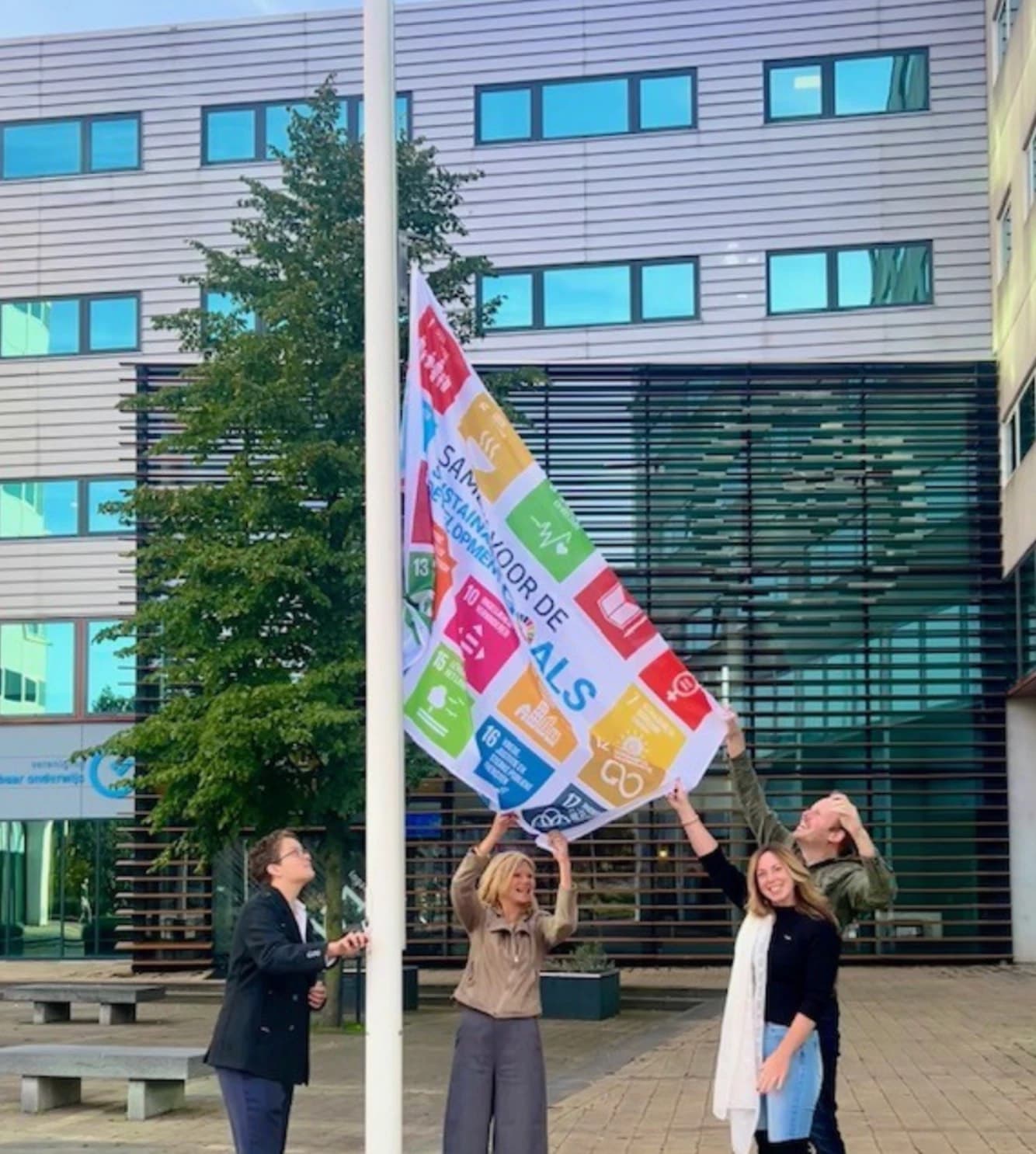 Vier mensen hijsen de vlag met de zeventien Duurzame Ontwikkelingsdoelen voor het gebouw van het OMO-bureau